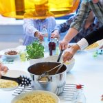 Mehrere Menschen die an einem Esstisch im freien Nudel aus einem Kochtopf schöpfen