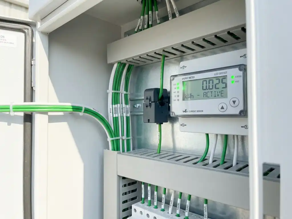 Digitaler Energiezähler mit Stromsensor im weißen Schaltschrank, grüne LED-Anzeigen, ordentlich verlegte Kabel.