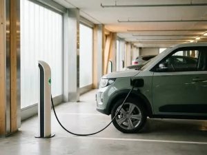 Elektro-Firmenwagen an weißer Ladestation in hellem Unternehmens-Parkhaus, grüne Statusanzeige leuchtet, Tageslicht durch große Fenster.