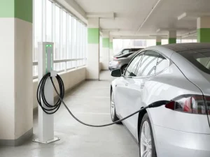 Elektroauto-Ladestation mit grünen LED-Anzeigen in hellem Firmenparkhaus, angeschlossenes Ladekabel, Tageslicht durch große Fenster.